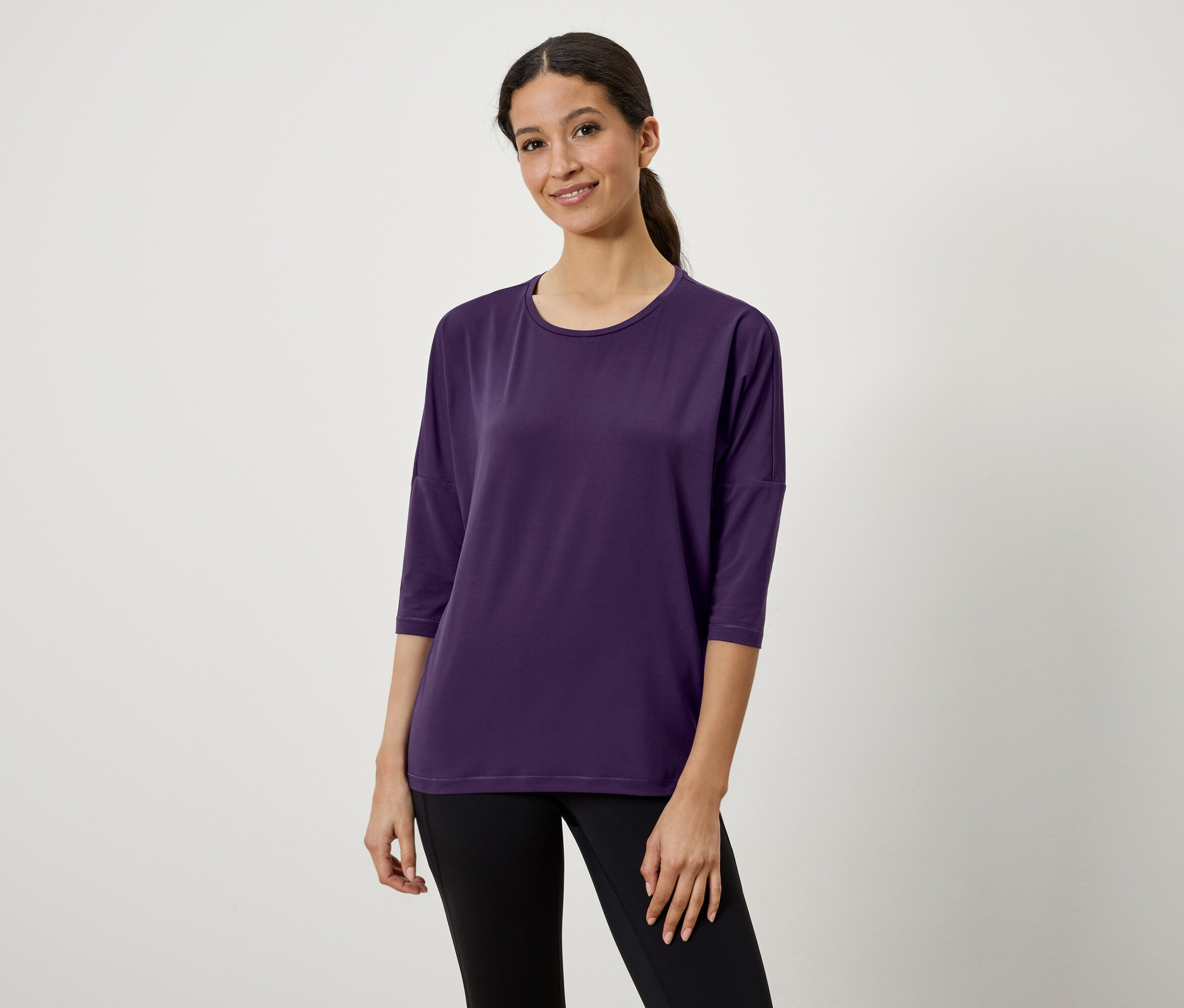 Žena s hnědými vlasy a culíkem má na sobě fialové tričko s tříčtvrtečními rukávy a černé legíny.