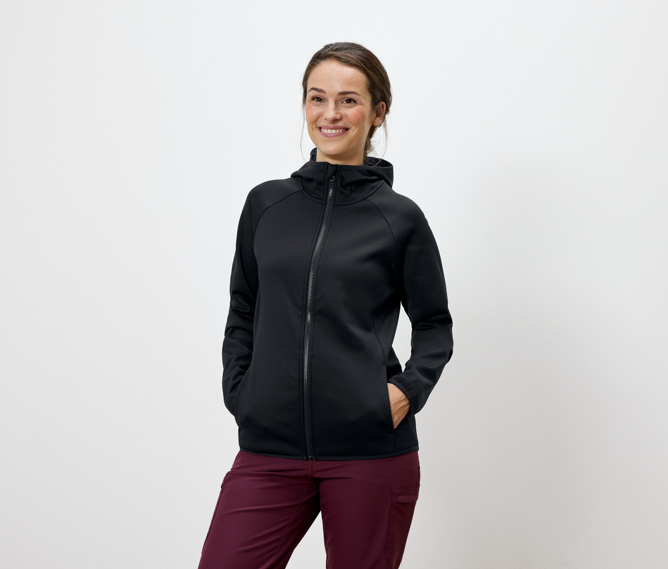 Žena pózuje v černé mikině s kapucí a vínových kalhotách.