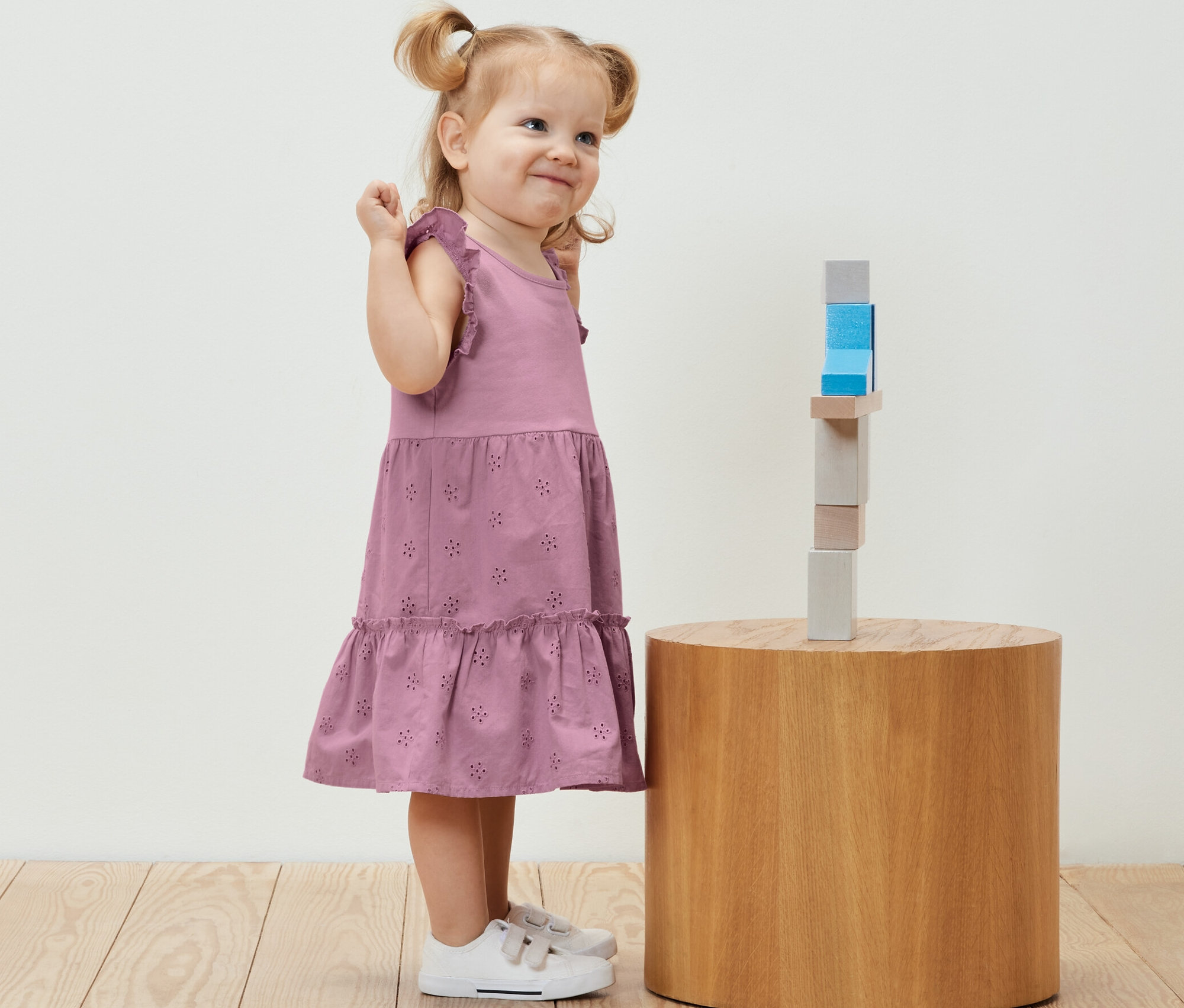 Usmívající se holčička v dětských šatech stojí vedle dřevěných kostek na kulatém stolku.