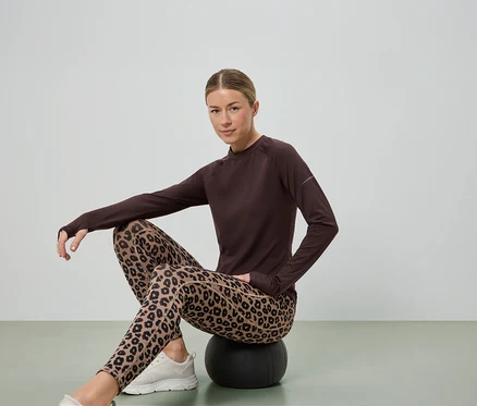 Žena sedí na černém míči, má na sobě hnědou blůzu s dlouhým rukávem, legíny s leopardím vzorem a bílé tenisky.