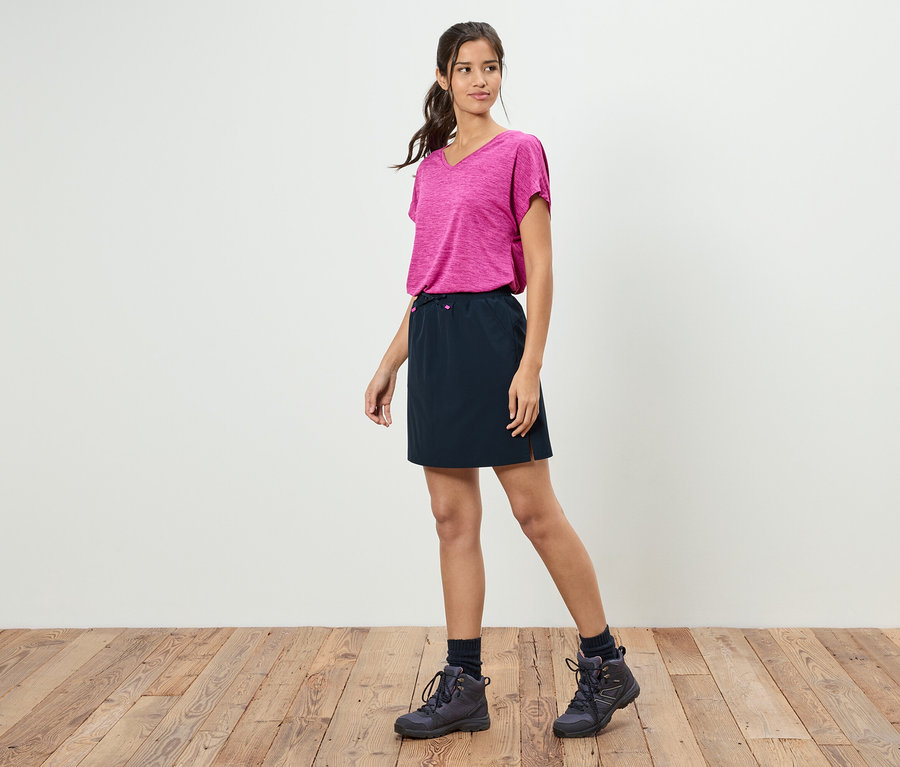 Žena stojí před bílou zdí a má na sobě růžové tričko, černou sukni a trekové boty.