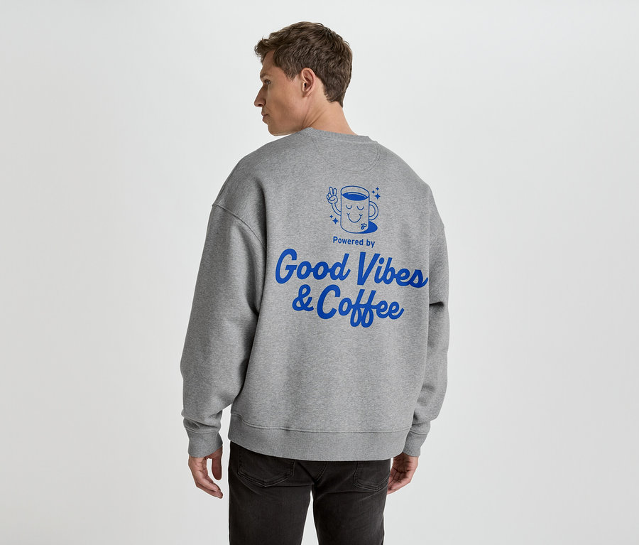 Muž zezadu v šedé mikině s modrým potiskem 'Good Vibes & Coffee'.