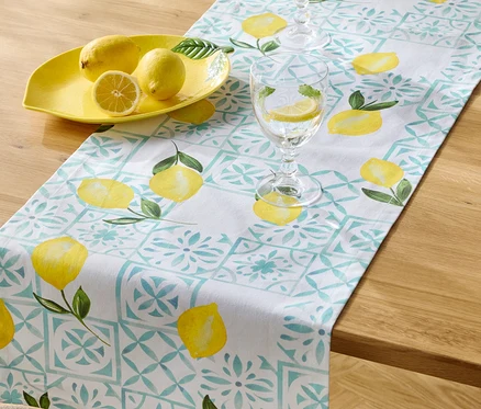 Na dřevěném stole leží běhoun se vzorem citronů, žlutá mísa na citrony se třemi citrony a dvě sklenice vody s plátky citronu.