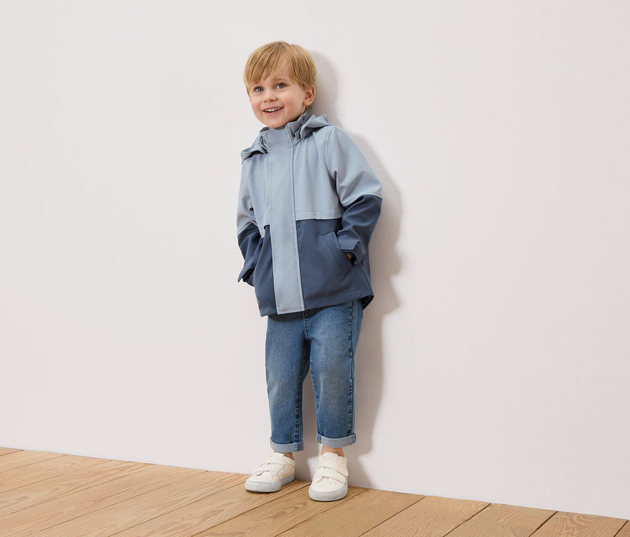 Usmívající se chlapec s blond vlasy stojí před bílou zdí a má na sobě dvoubarevnou, světle modrou a námořnickou bundu, modré džíny a bílé tenisky.