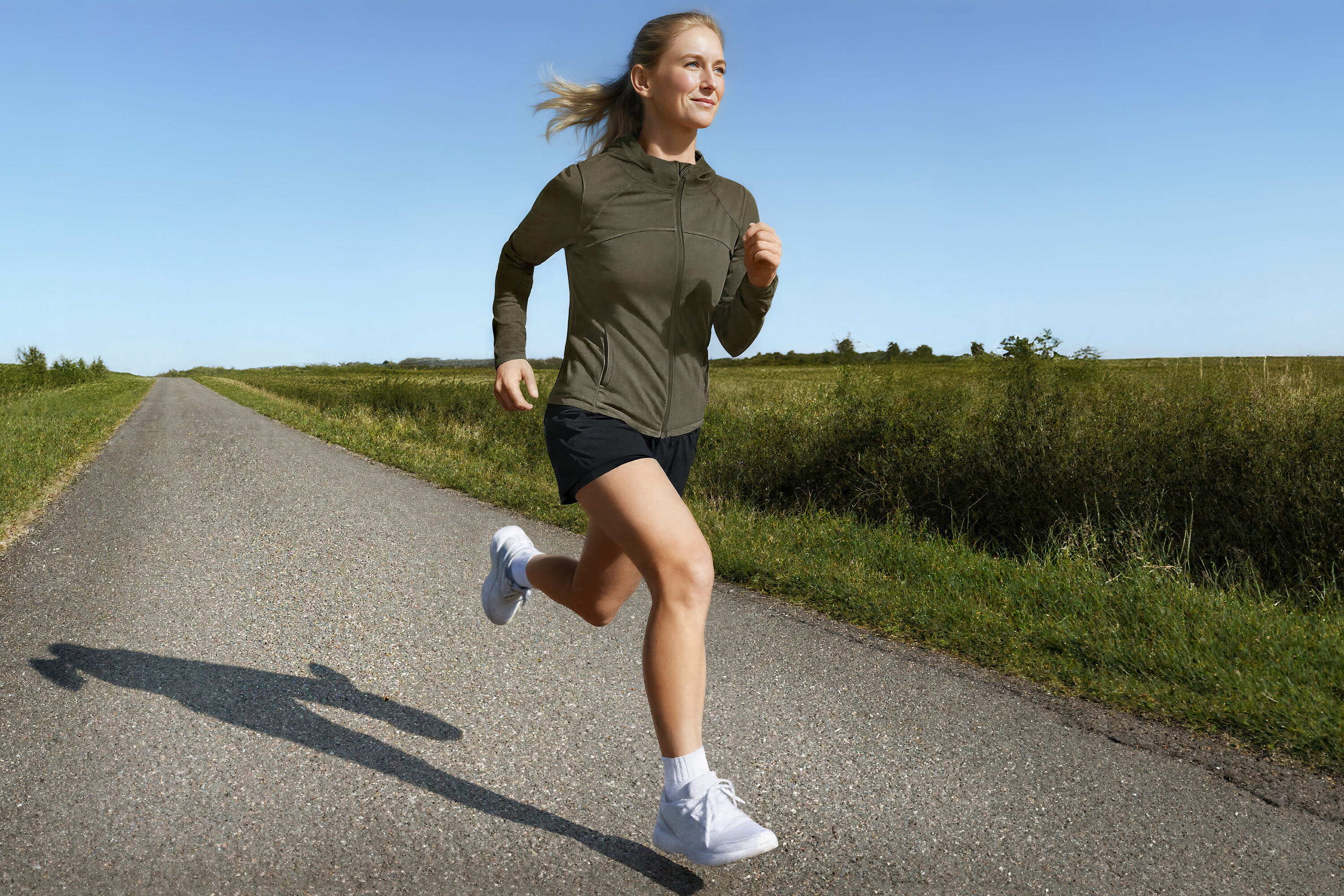 Žena běží po venkovské silnici ve sportovní bundě a běžeckých šortkách 2 v 1.