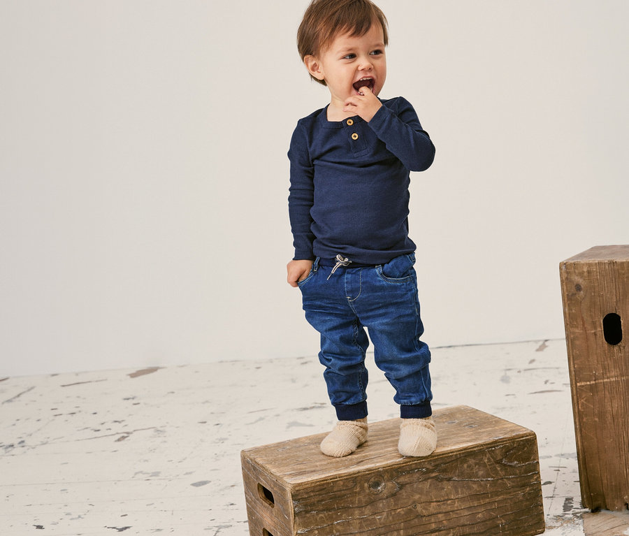 Batole stojí na dřevěné bedně a má na sobě dětské žebrované triko s dlouhým rukávem NAME IT v tmavě modré barvě.