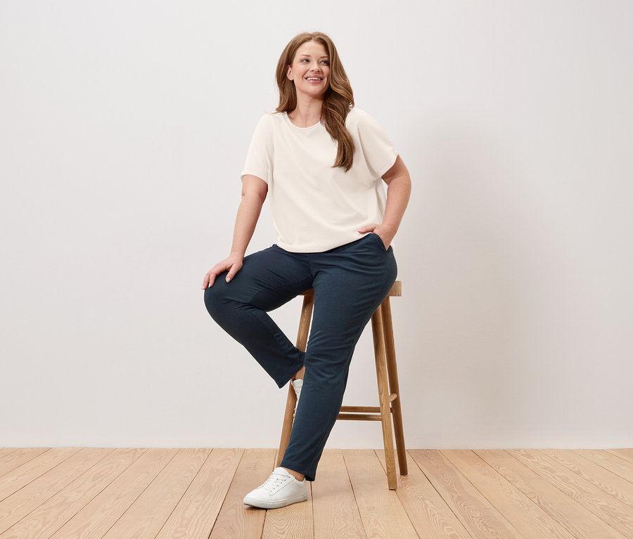 Žena sedí na stoličce, má na sobě krémové tričko, modré kalhoty a bílé tenisky.