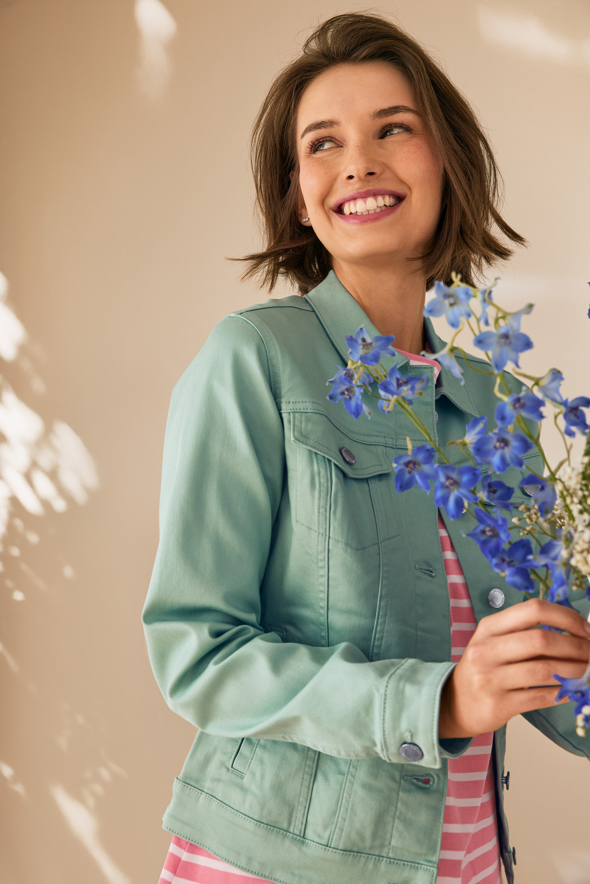 Žena se usmívá a drží modré květiny, má na sobě mátově zelenou džínovou bundu Džínová bunda, mátová zelená.