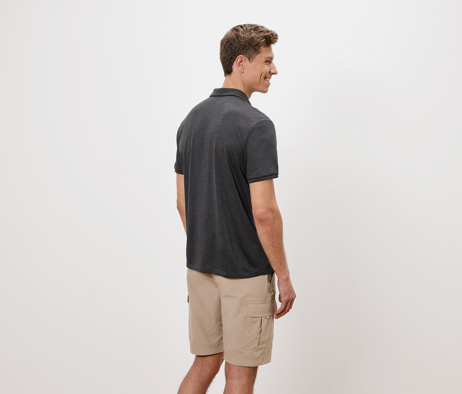 Muž stojí bokem, má na sobě šedé polo tričko a béžové cargo šortky na bílém pozadí.