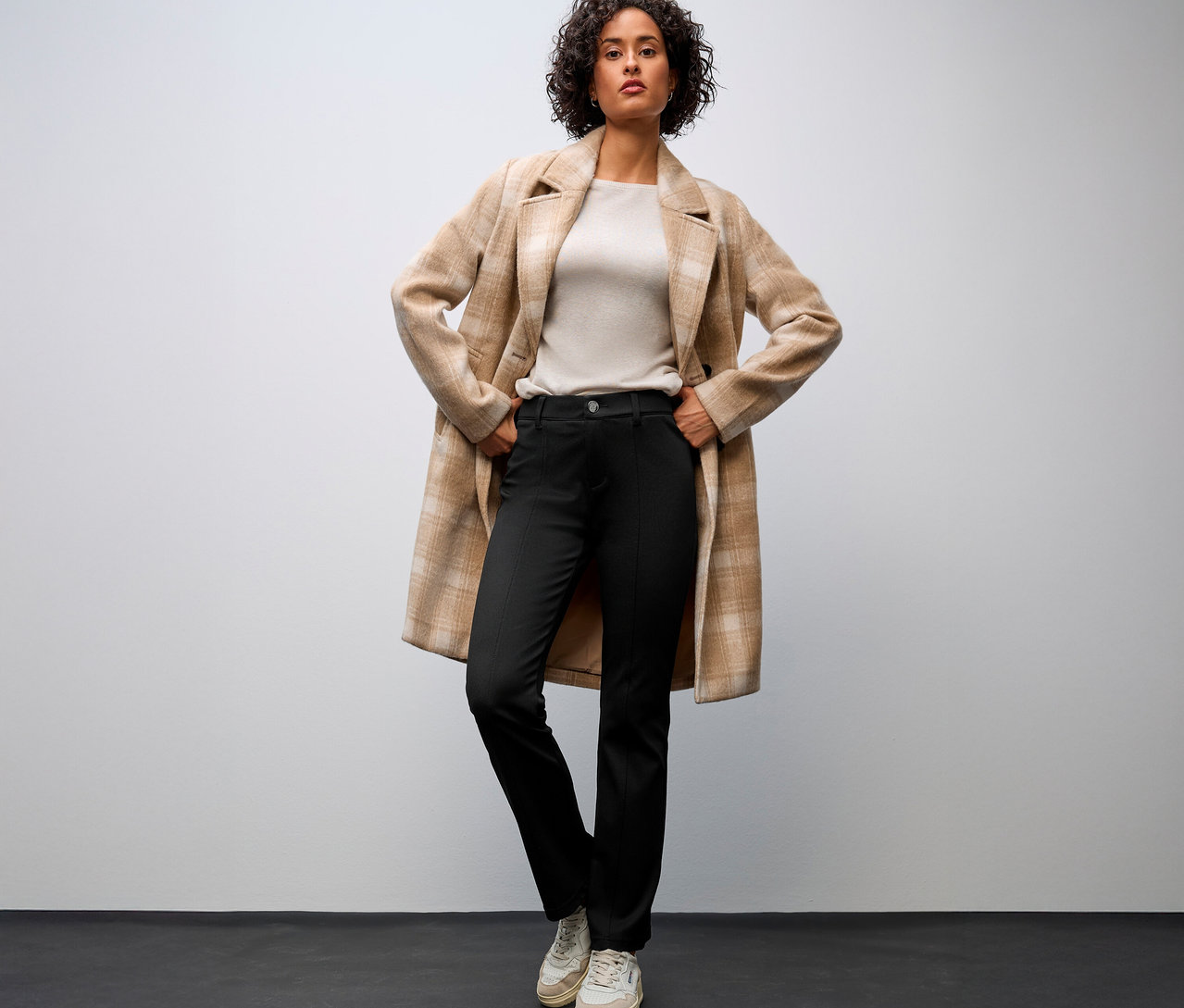 Žena v béžovém kabátě a kalhotách s rozšířenými nohavicemi Street One s rukama v bocích.