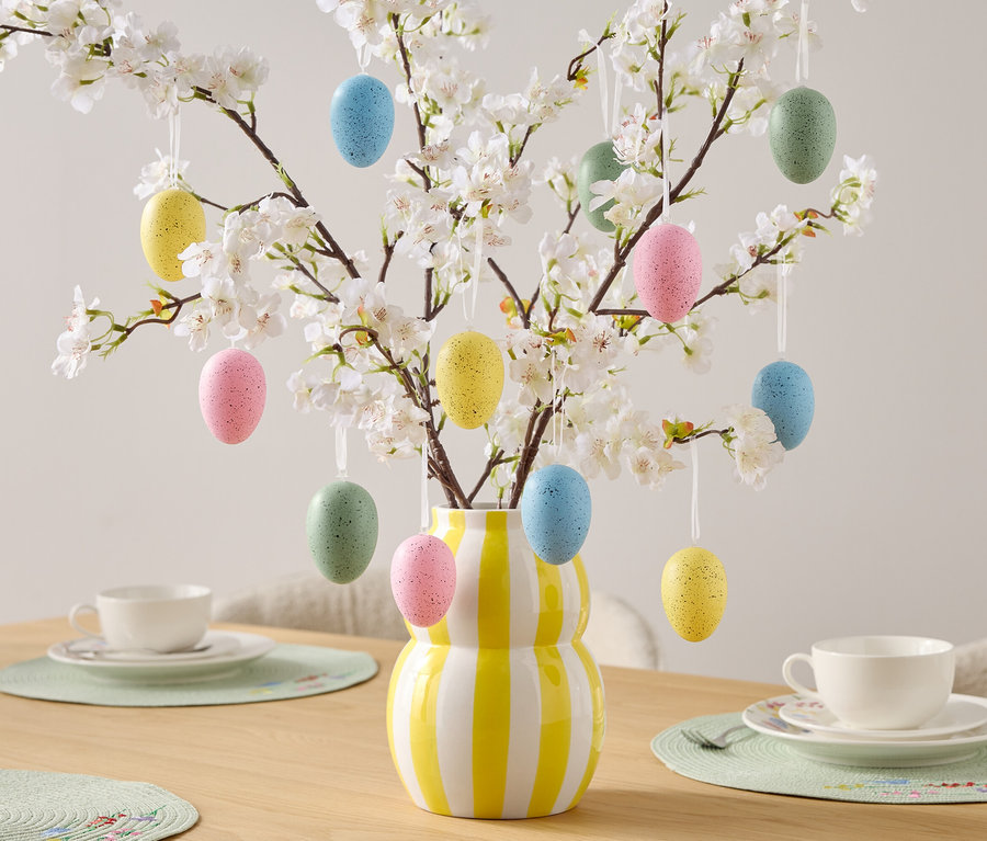 Dekorativní velikonoční vajíčka visí na kvetoucích větvích ve váze se žluto-bílými pruhy, stojící na stole s prostíráním.