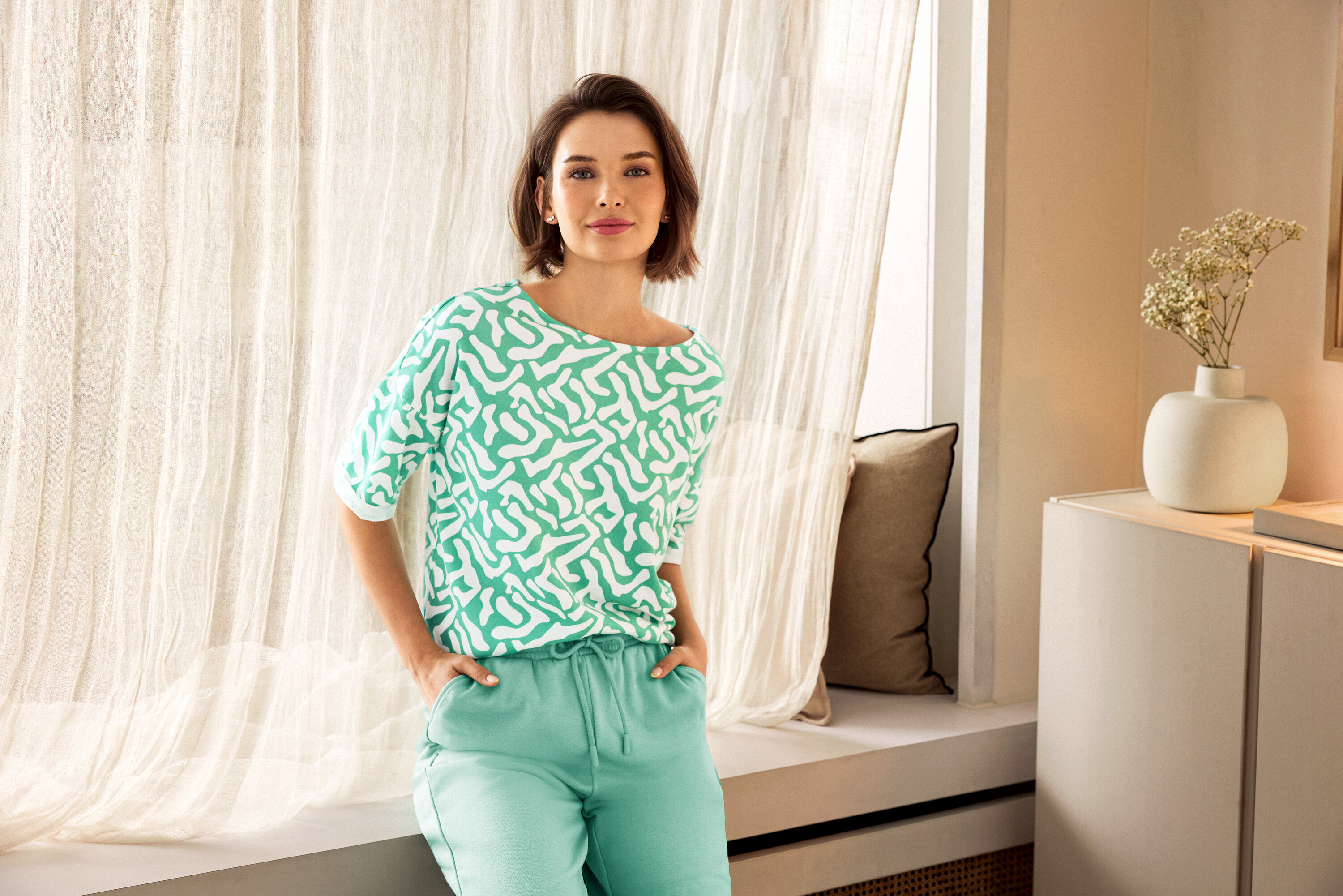Žena v zelených kalhotách a vzorované halence pózuje u okna. V pozadí je vidět váza.