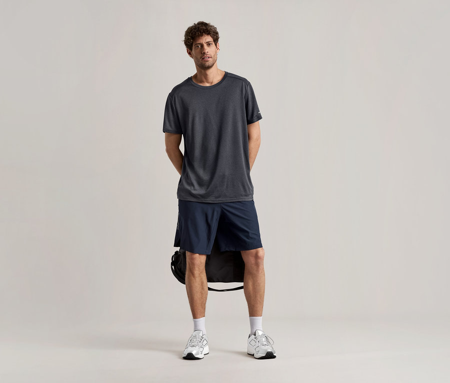 Muž stojí na světlém pozadí ve sportovním tričku antracitové barvy, v šortkách a teniskách.