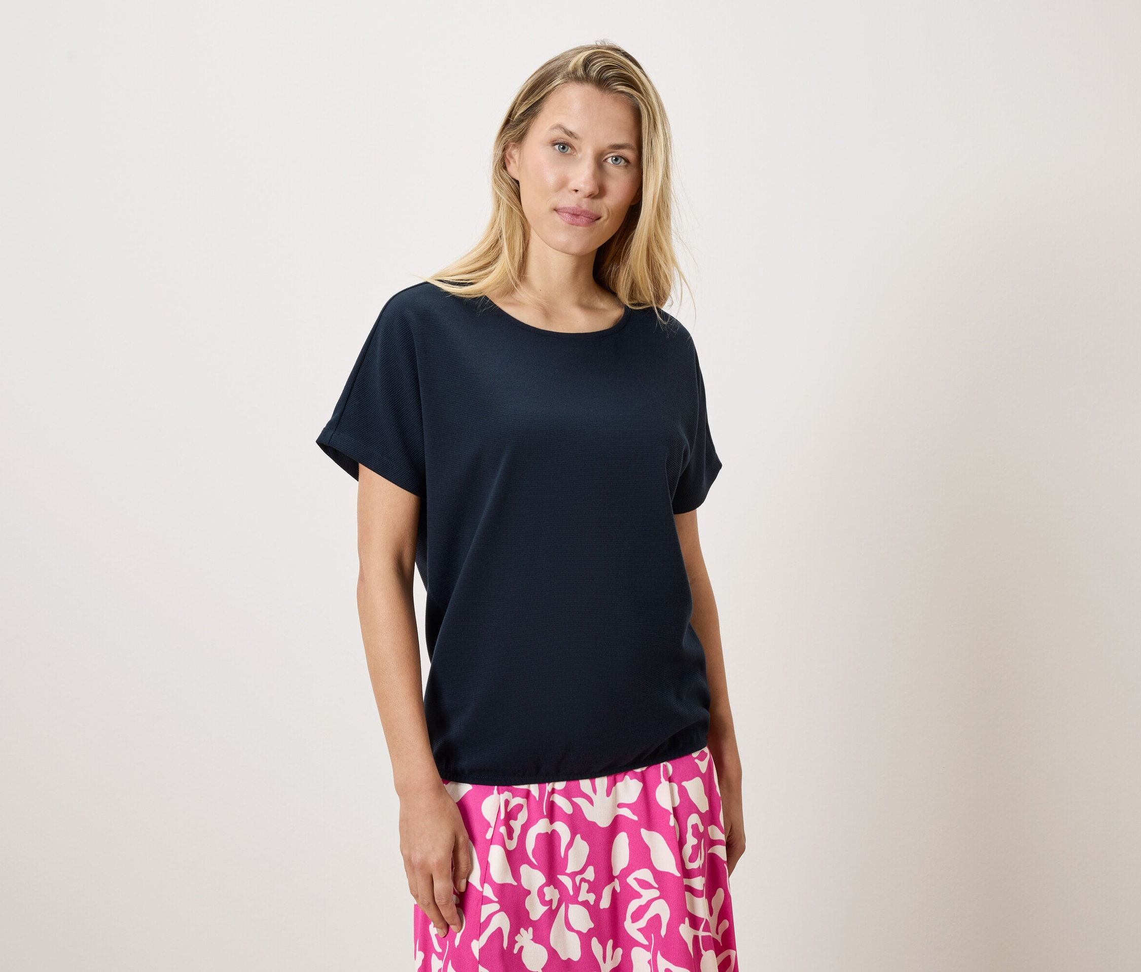 Blondýna má na sobě tmavě modré tričko a růžovo-bílou sukni na bílém pozadí.