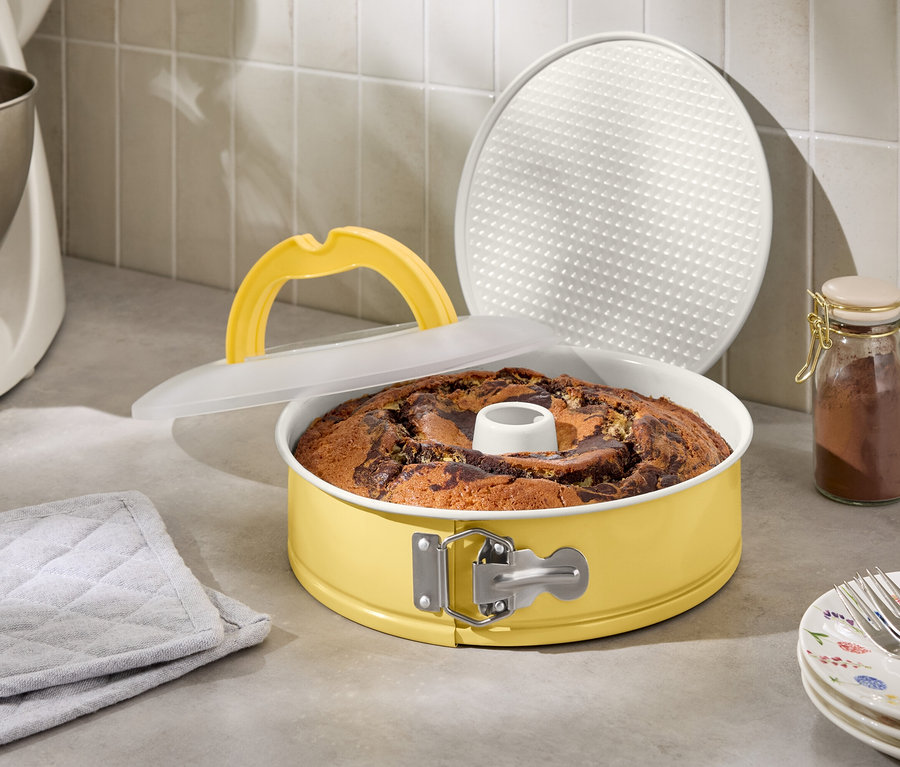 Mramorová bábovka ve žluté dortové formě s víkem a zvedací rukojetí vedle kuchyňského robota a sklenice s kořením