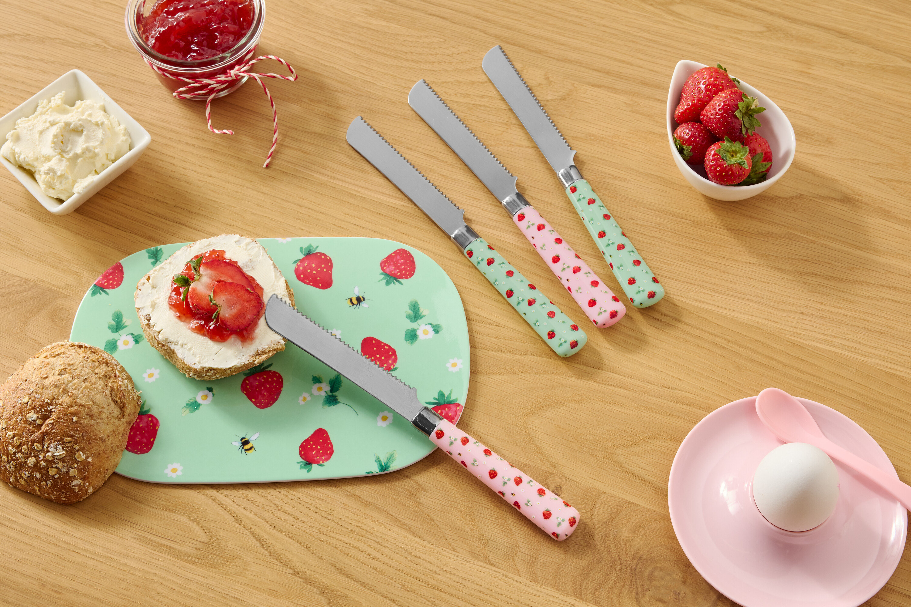 Na dřevěném stole je houska, talíř se vzorem jahod, smetanový sýr, džem, jahody a čtyři nože se vzorem jahod.