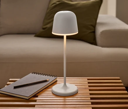 Svítící bílá LED dobíjecí stolní lampa Eglo »Mannera« stojí na lamelovém dřevěném stole vedle zápisníku s tužkou před pohovkou.