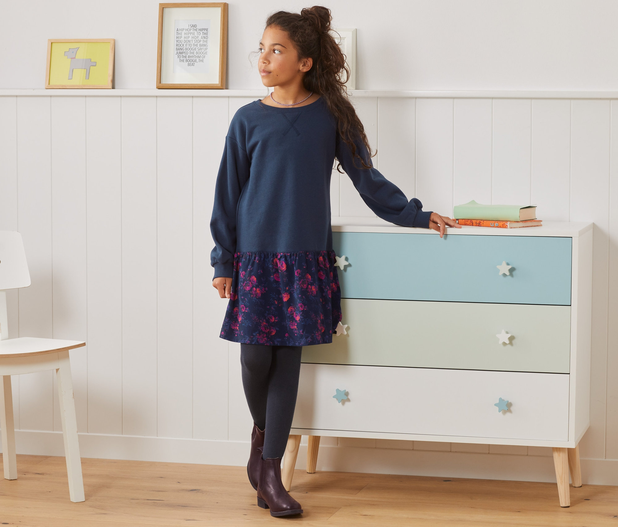 Dívka stojí a opírá se o komodu v teplákových šatech.