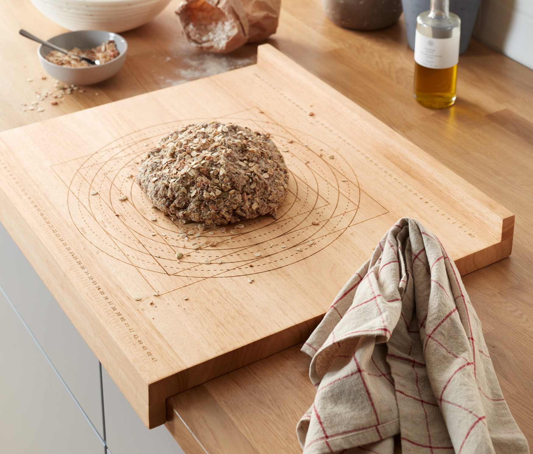 Na kuchyňské lince leží dřevěný kuchyňský vál na těsto s bochníkem chleba. V pozadí jsou bylinky v květináčích a další kuchyňské potřeby.