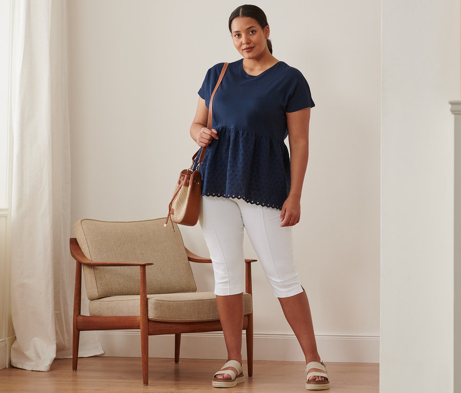 Žena stojí a má na sobě tmavě modré triko z kombinace materiálů s výšivkou, 3/4 strečové kalhoty, kožené sandály na platformě a kabelku.