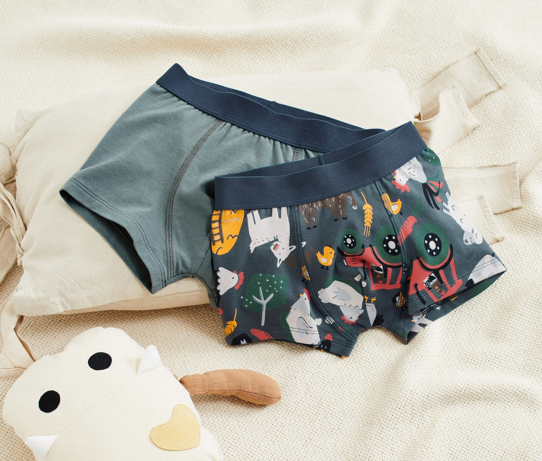 Dvoje chlapecké boxerky leží na béžové látce vedle plyšové hračky.