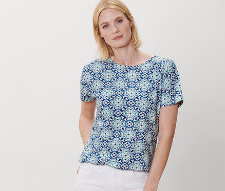 Žena pózuje v halénkovém tričku s celoplošným potiskem a bílých kalhotách.