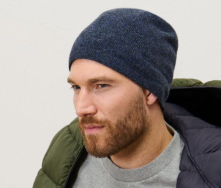 Muž má na sobě pletenou čepici tmavě modré barvy s melírem, šedivý svetr a zelenou péřovou bundu.