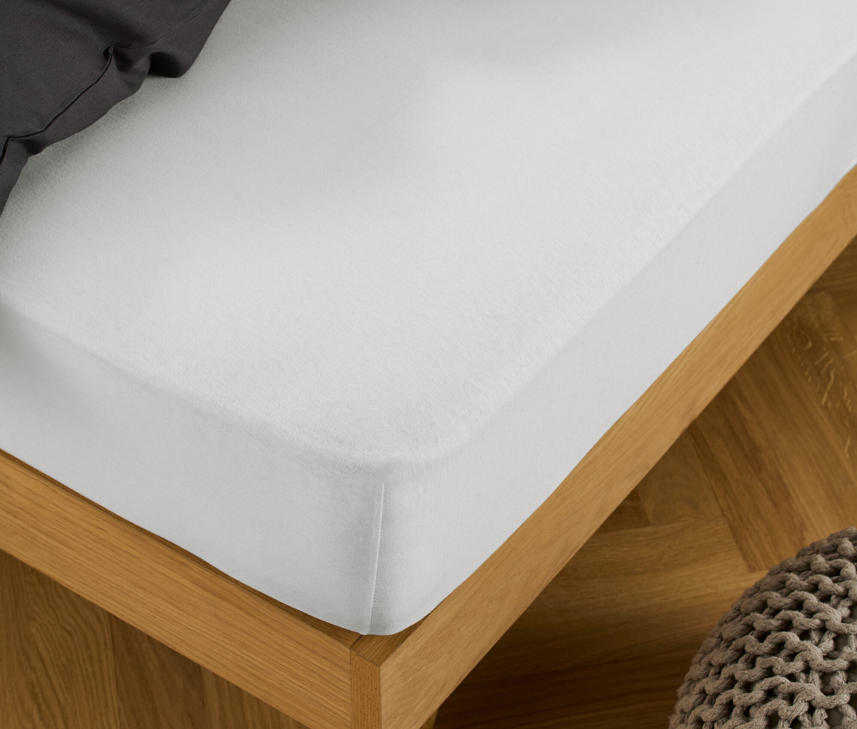 Bílé žerzejové napínací prostěradlo je natažené na posteli s dřevěným rámem, vedle stojí pletený puf.
