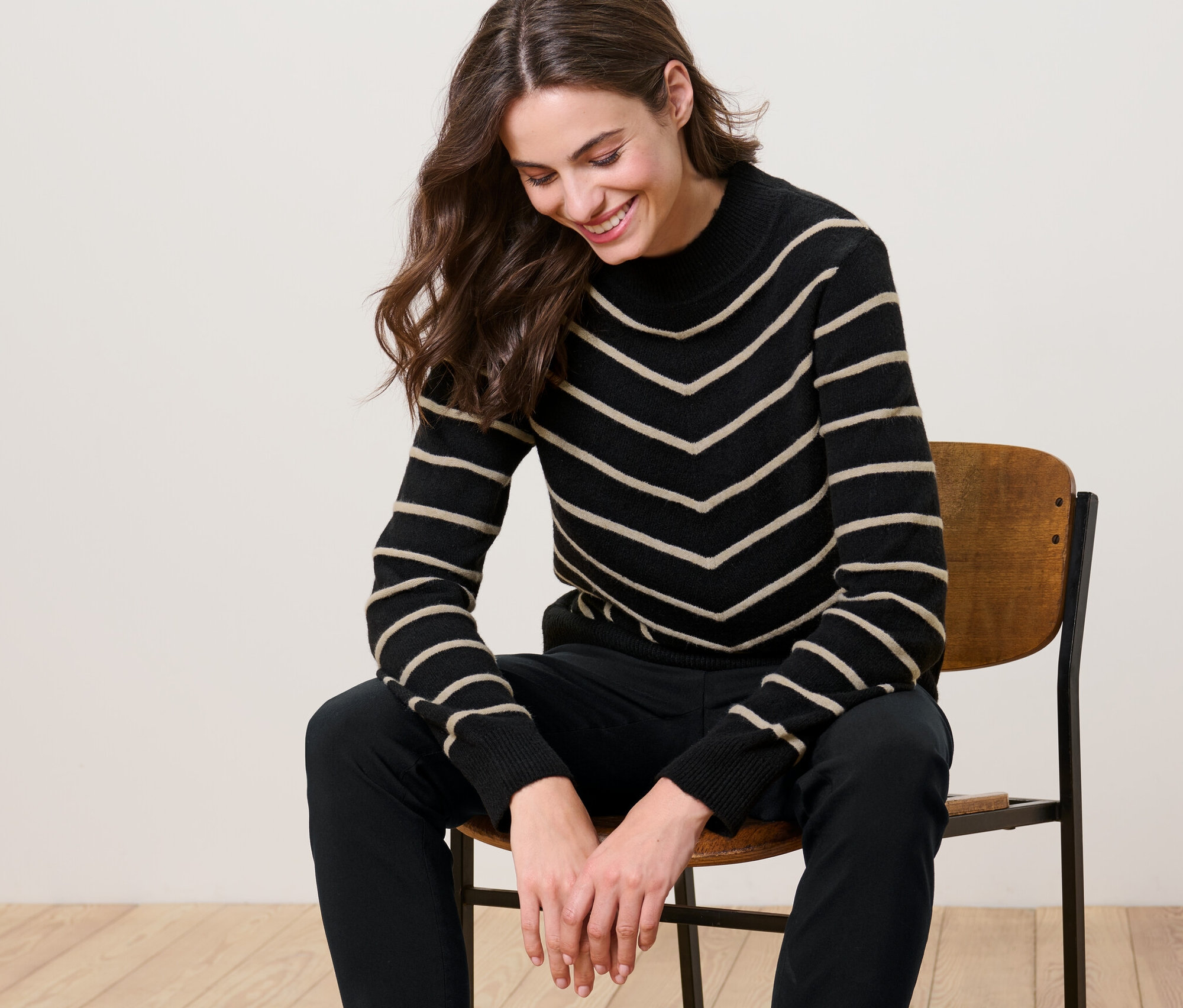 Žena sedí na židli a má na sobě černý pletený svetr se stojáčkem a proužky.