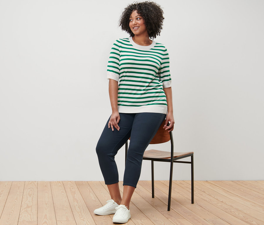 Žena sedí na židli a má na sobě proužkovaný svetr z jemného úpletu a 7/8 džegíny.