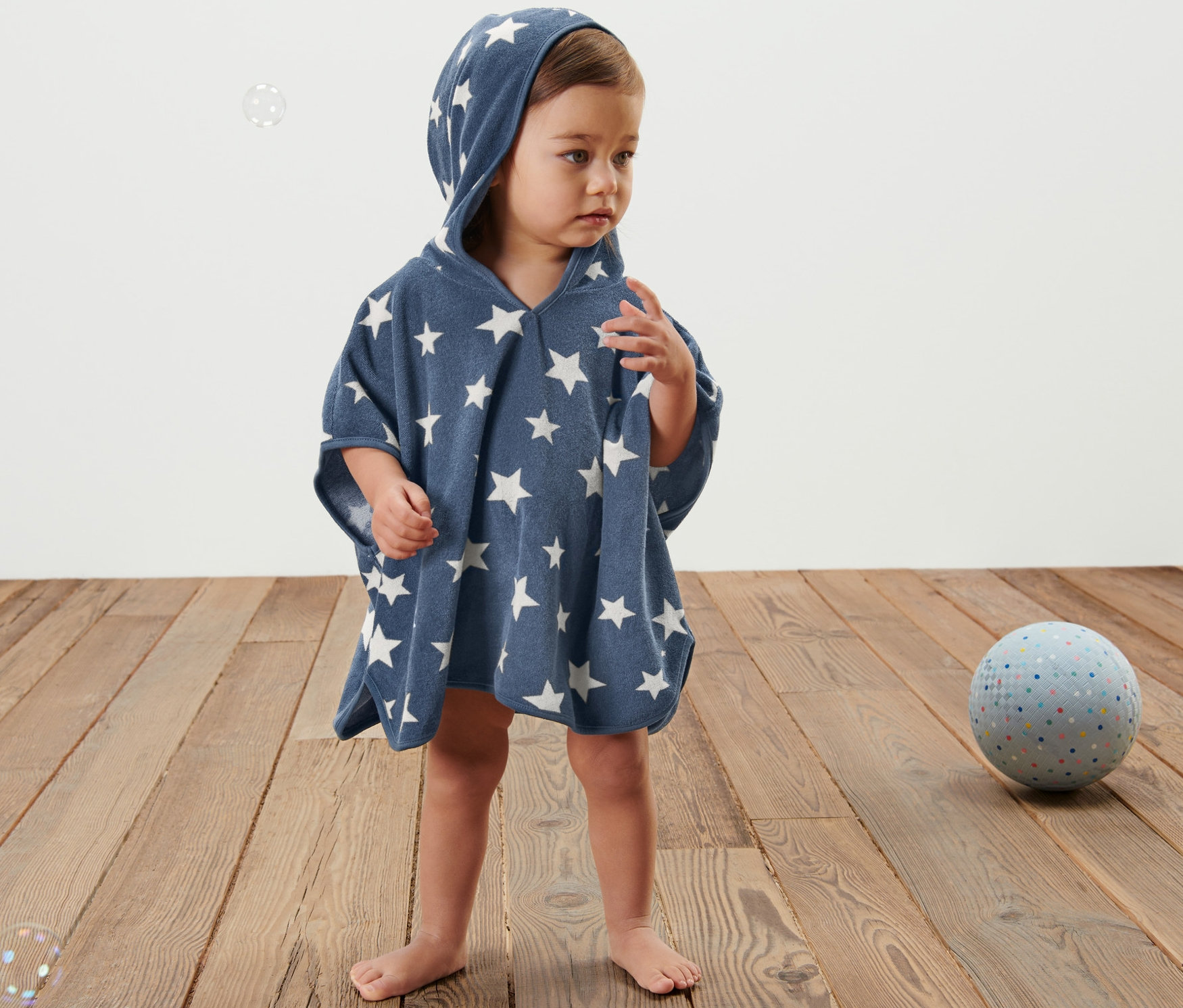 Dítě stojí na dřevěné podlaze a má na sobě modré froté pončo s kapucí a bílými hvězdami.