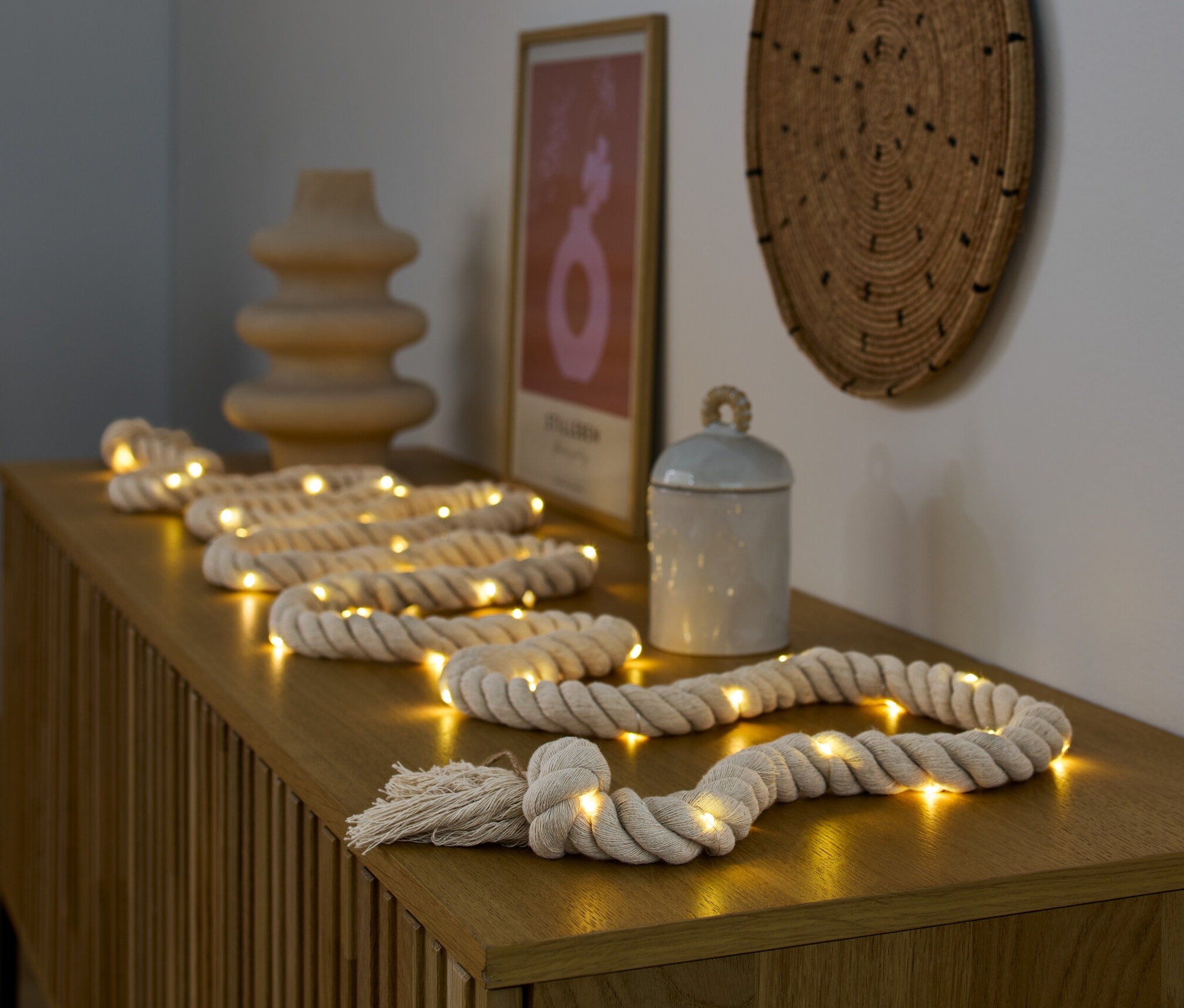LED světelný řetěz »Lano« leží na dřevěné komodě vedle vázy a dózy.