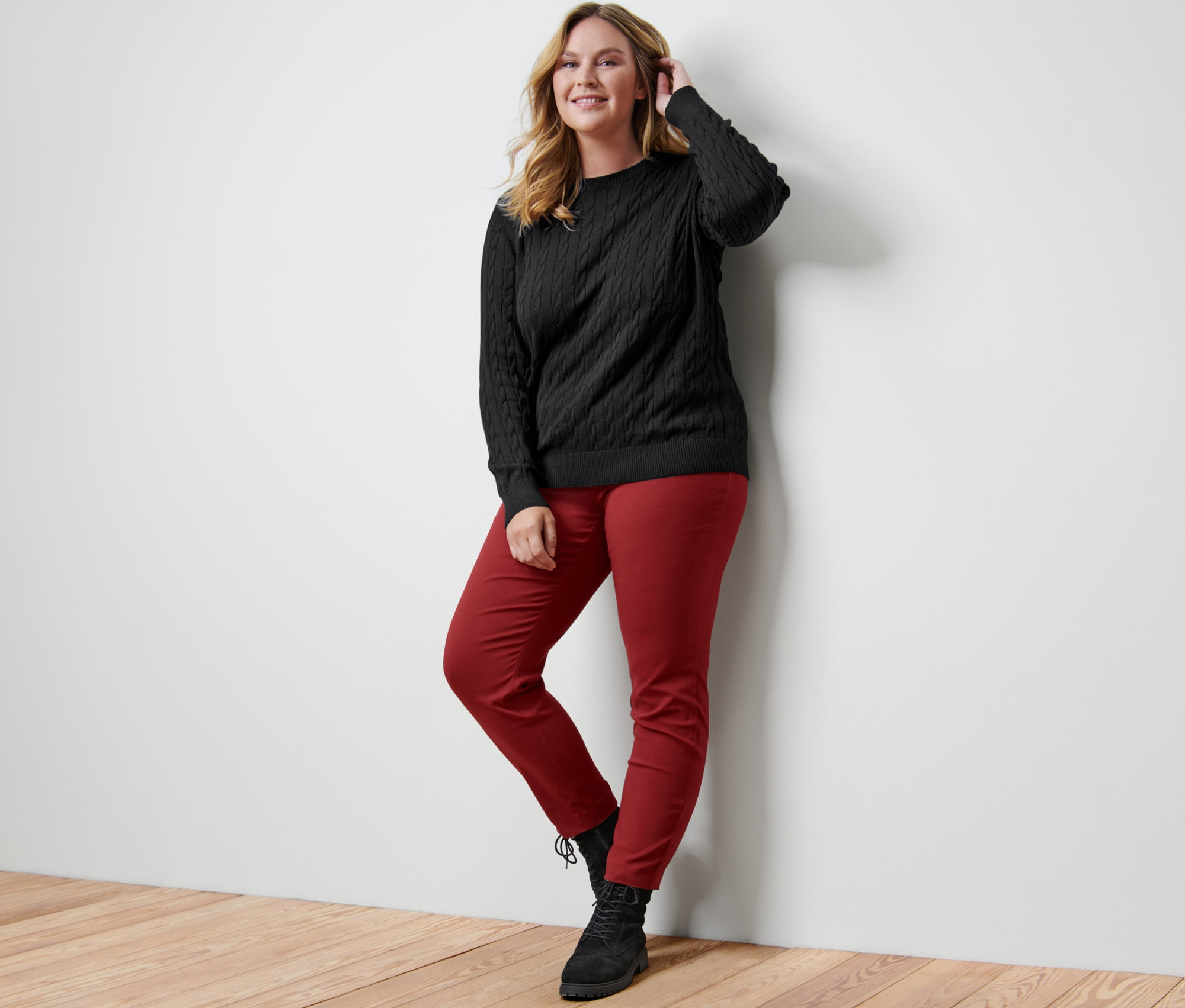 Žena pózuje opřená o zeď a má na sobě černý svetr s copánkovým vzorem, červené strečové kalhoty a kotníkové boty z velurové kůže.