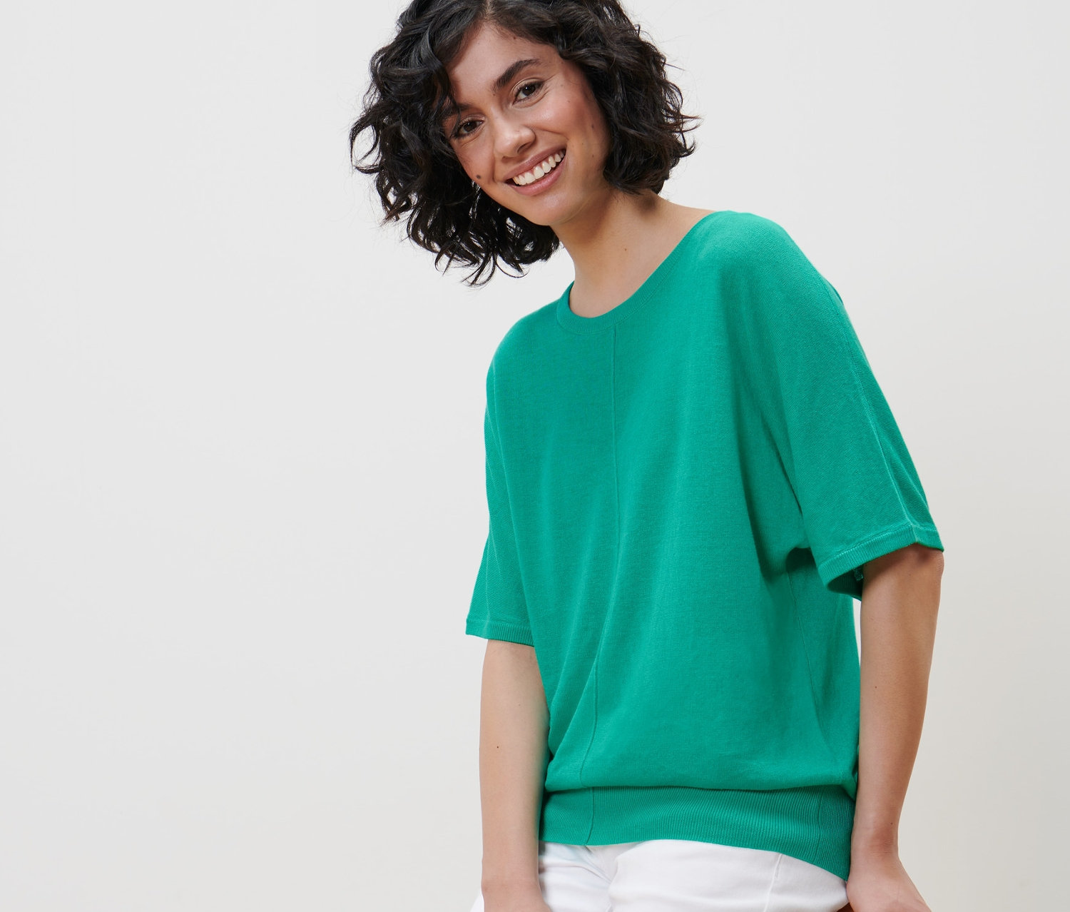 Žena sedí na židli a má na sobě zelené tričko z jemného úpletu s lnem a bílé kalhoty.