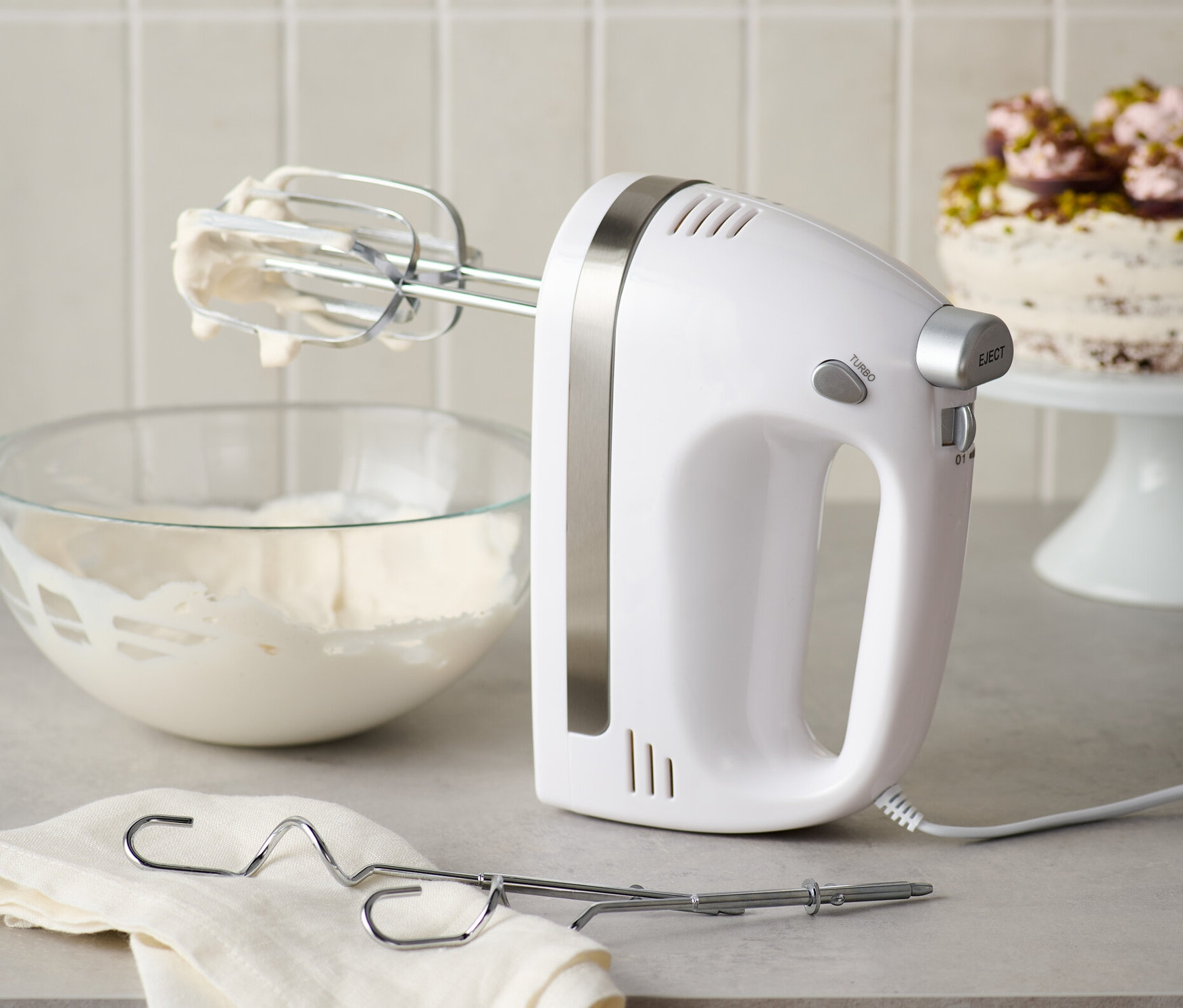 Bílý ruční mixér s nerezovými metlami stojí vedle mísy se šlehačkou a hadříku.