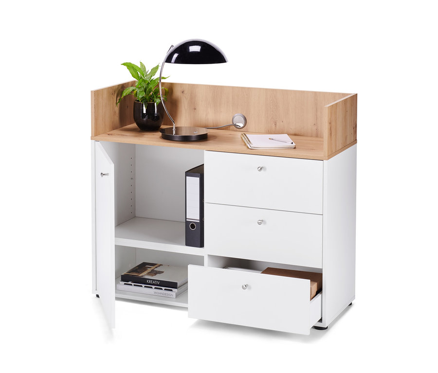 Bílá komoda se zásuvkami a dvířky, na níž stojí lampa a rostlina.