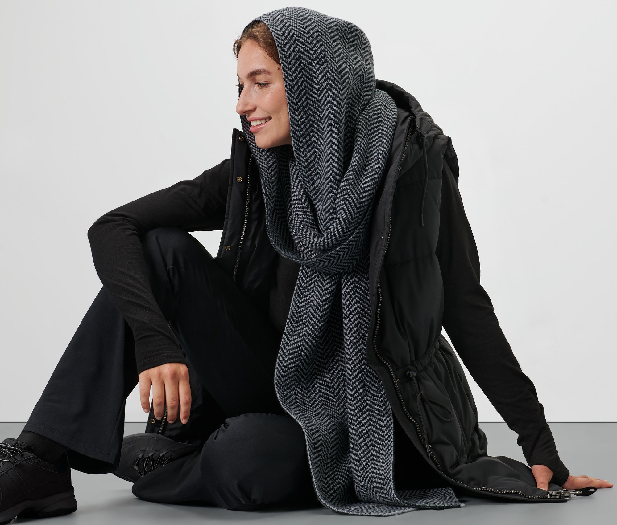 Žena sedí na podlaze a má na sobě černou vestu a šedou kapucovou šálu.