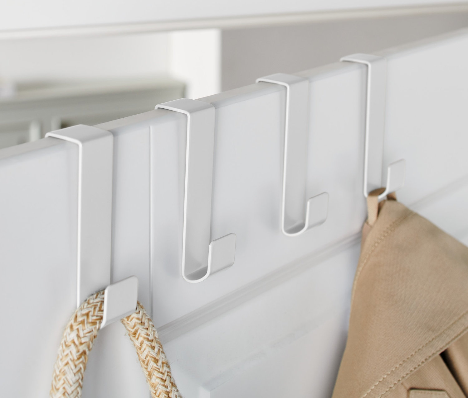 Na bílých dveřích jsou zavěšeny tři bílé háčky na dveře, na kterých visí bunda a taška.