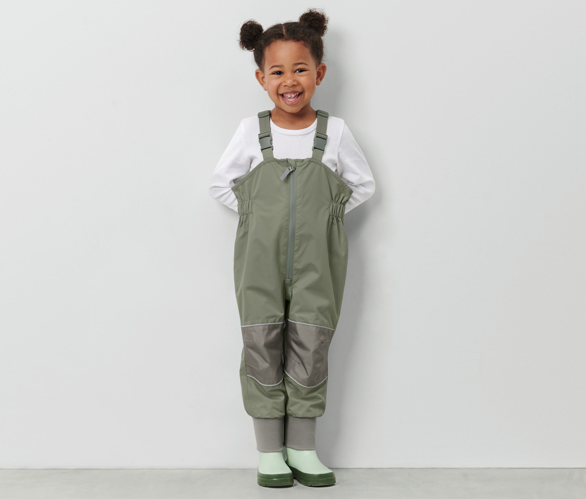 Usmívající se holčička stojí a má na sobě zelené dětské kalhoty do deště a zelené holínky.