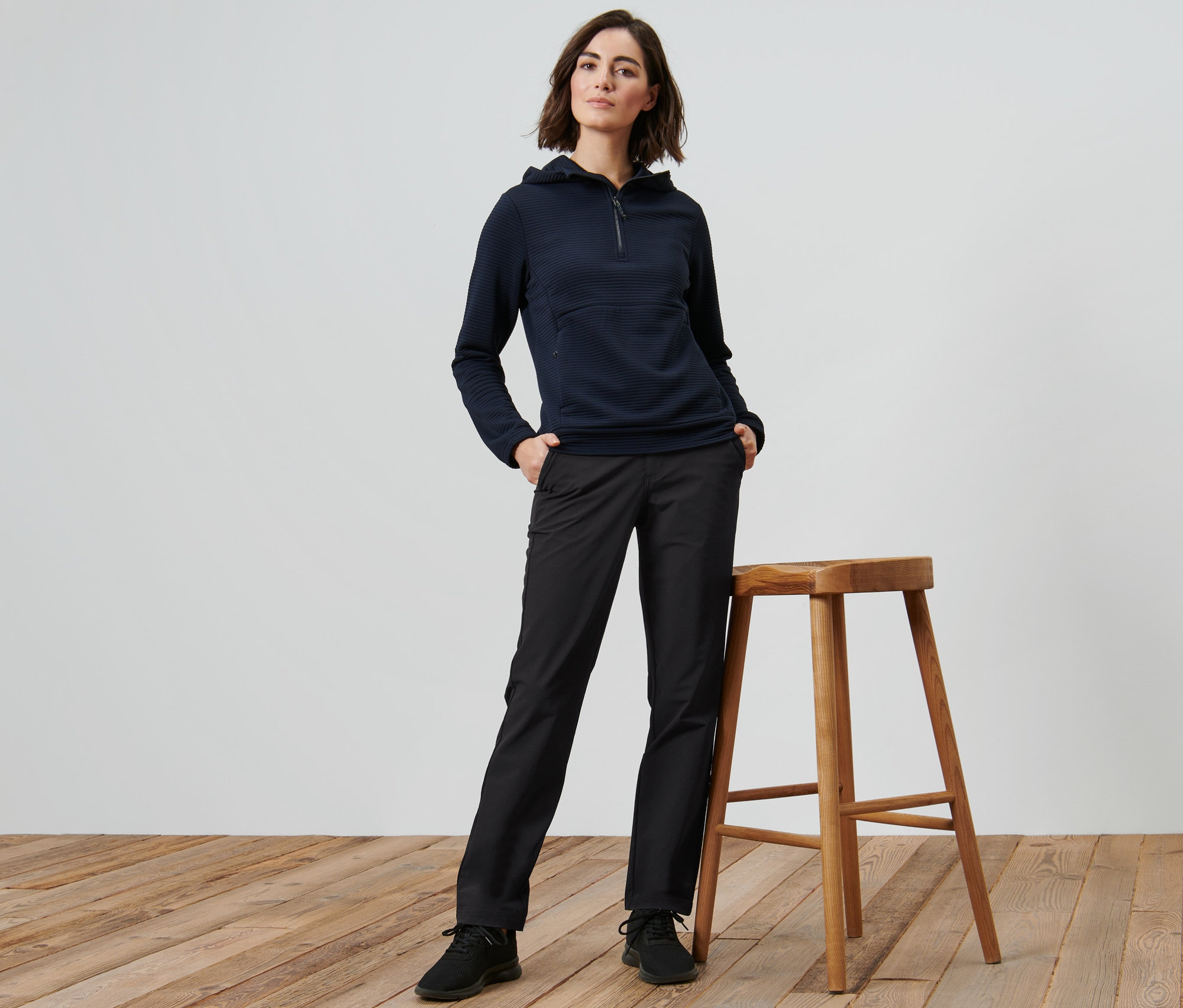 Žena pózuje vedle dřevěné stoličky a má na sobě softshellové kalhoty.