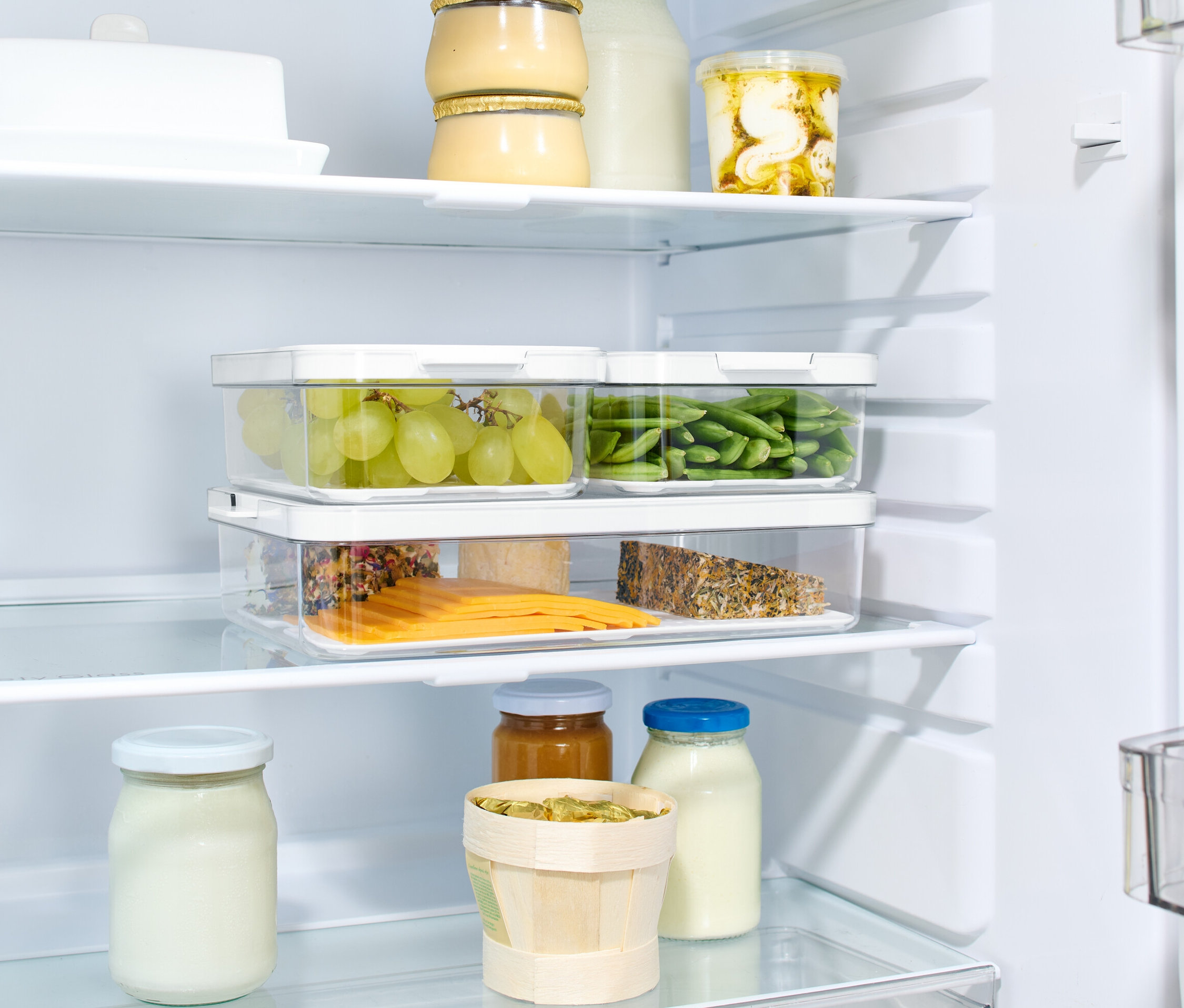 V lednici jsou tři úložné boxy s víkem. V boxech jsou zelené hrozny, zelené fazolky a plátky sýra.