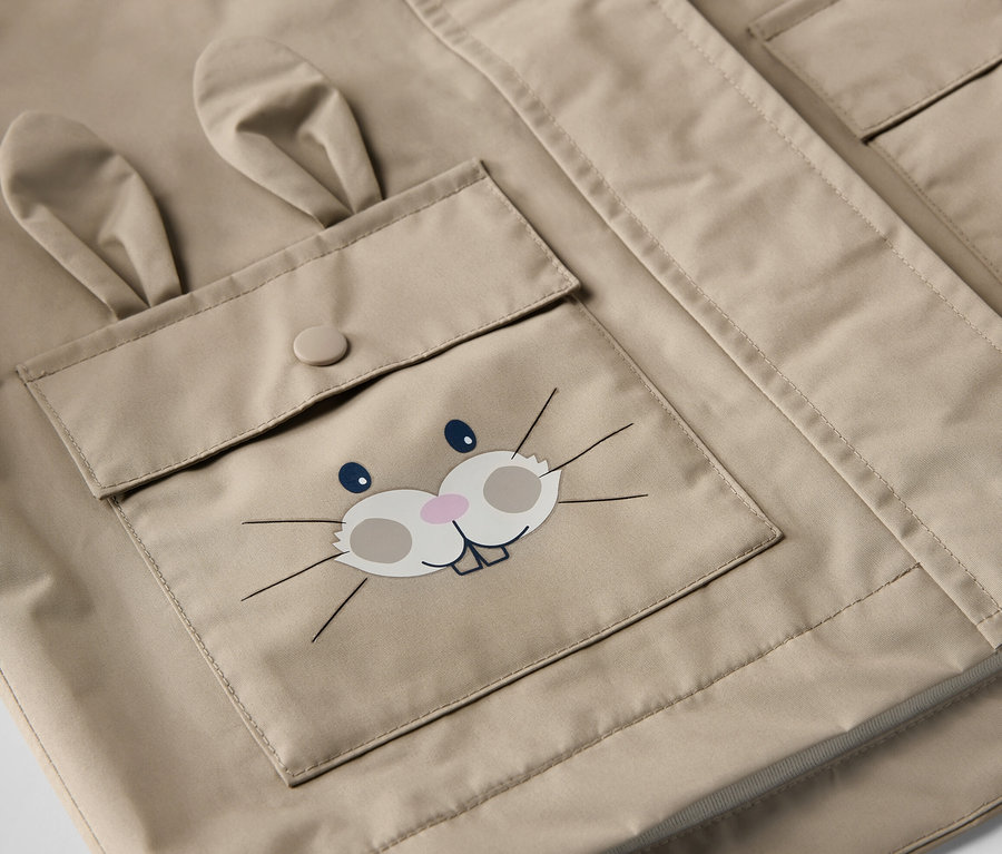 Detail dětské hnědé bundy do deště s kapsou s obličejem králíka.