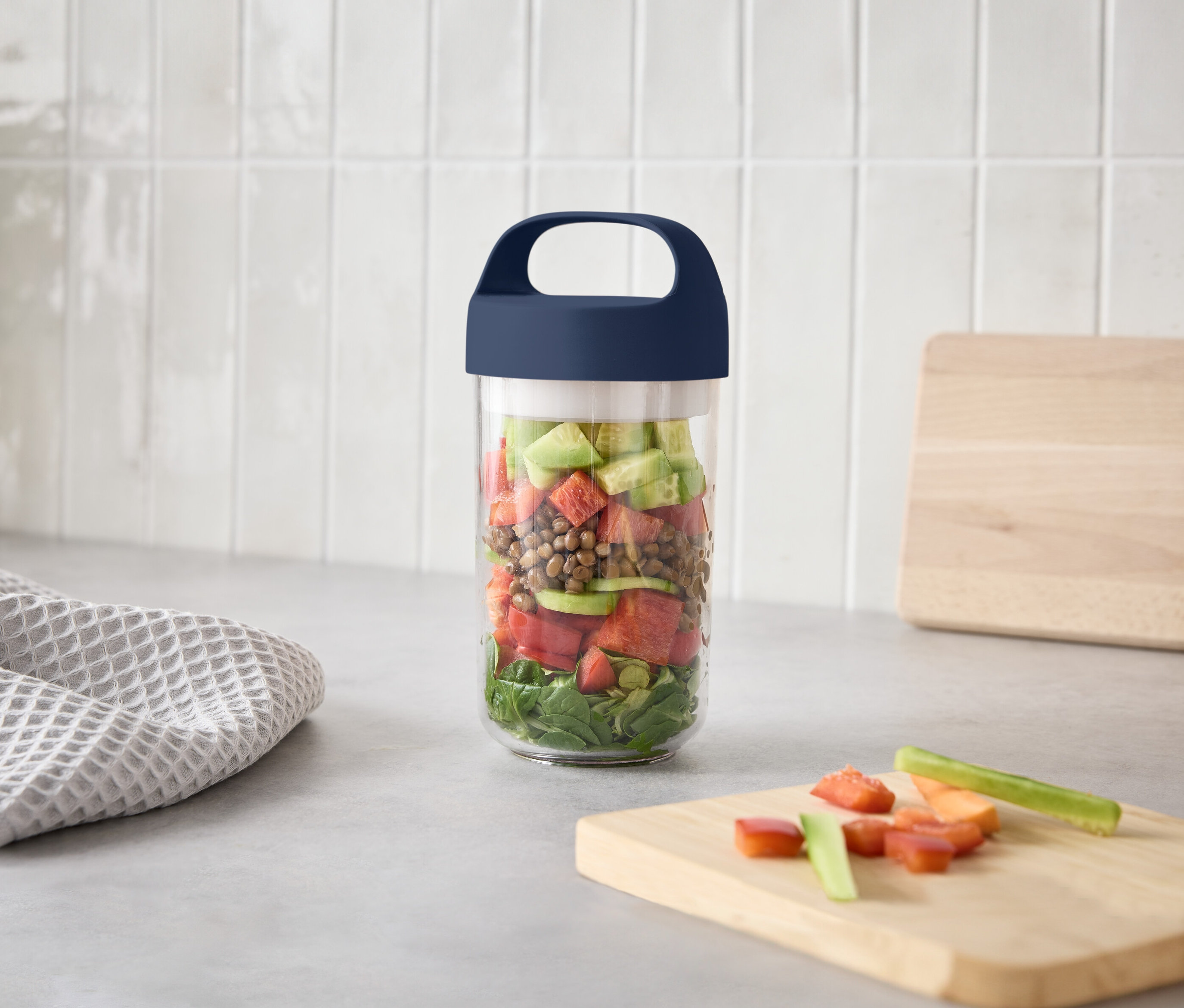 Sklenice s vrstvami salátu na kuchyňské lince vedle prkénka s nakrájenou zeleninou.