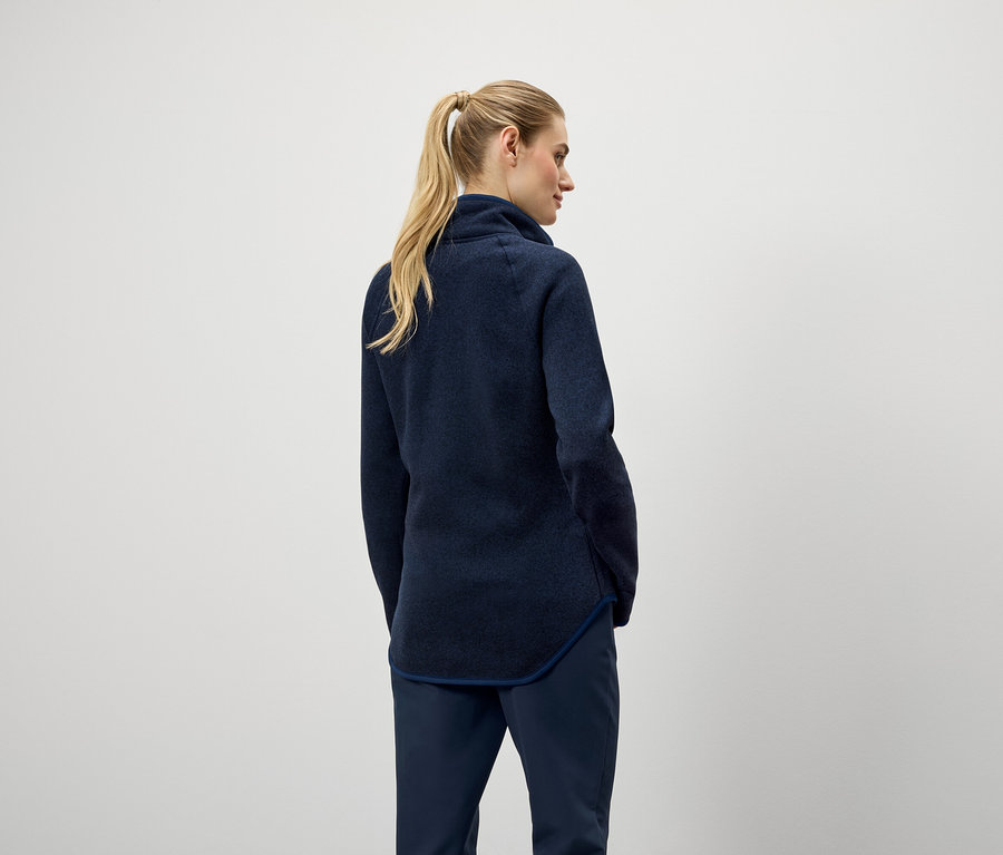 Zezadu je vidět žena s blond vlasy staženými do culíku, oblečená v tmavě modrém svetru a kalhotách.