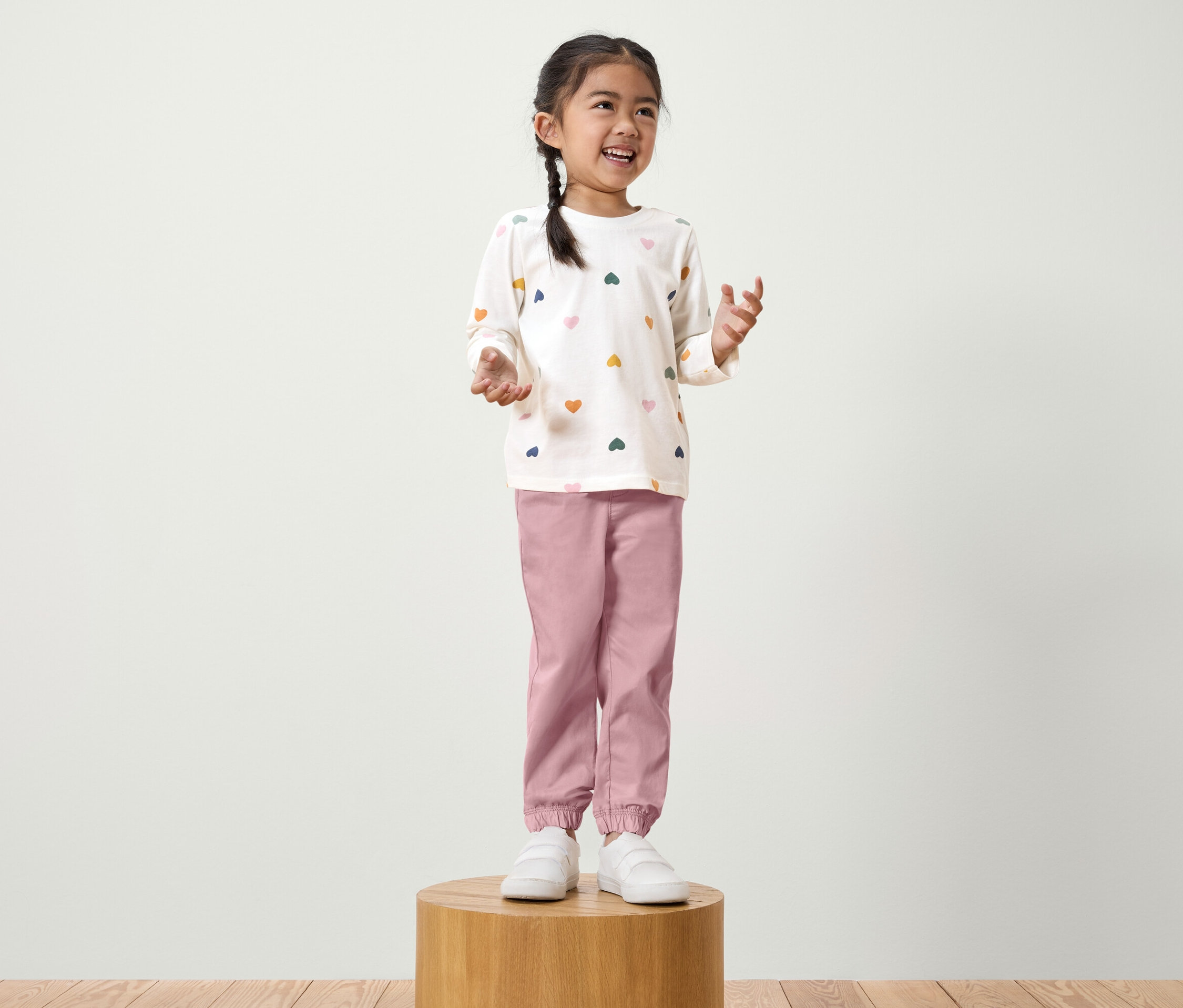 Dívka s copem stojí na dřevěném podstavci. Má na sobě bílé tričko s dlouhým rukávem s barevnými srdíčky a růžové kalhoty.
