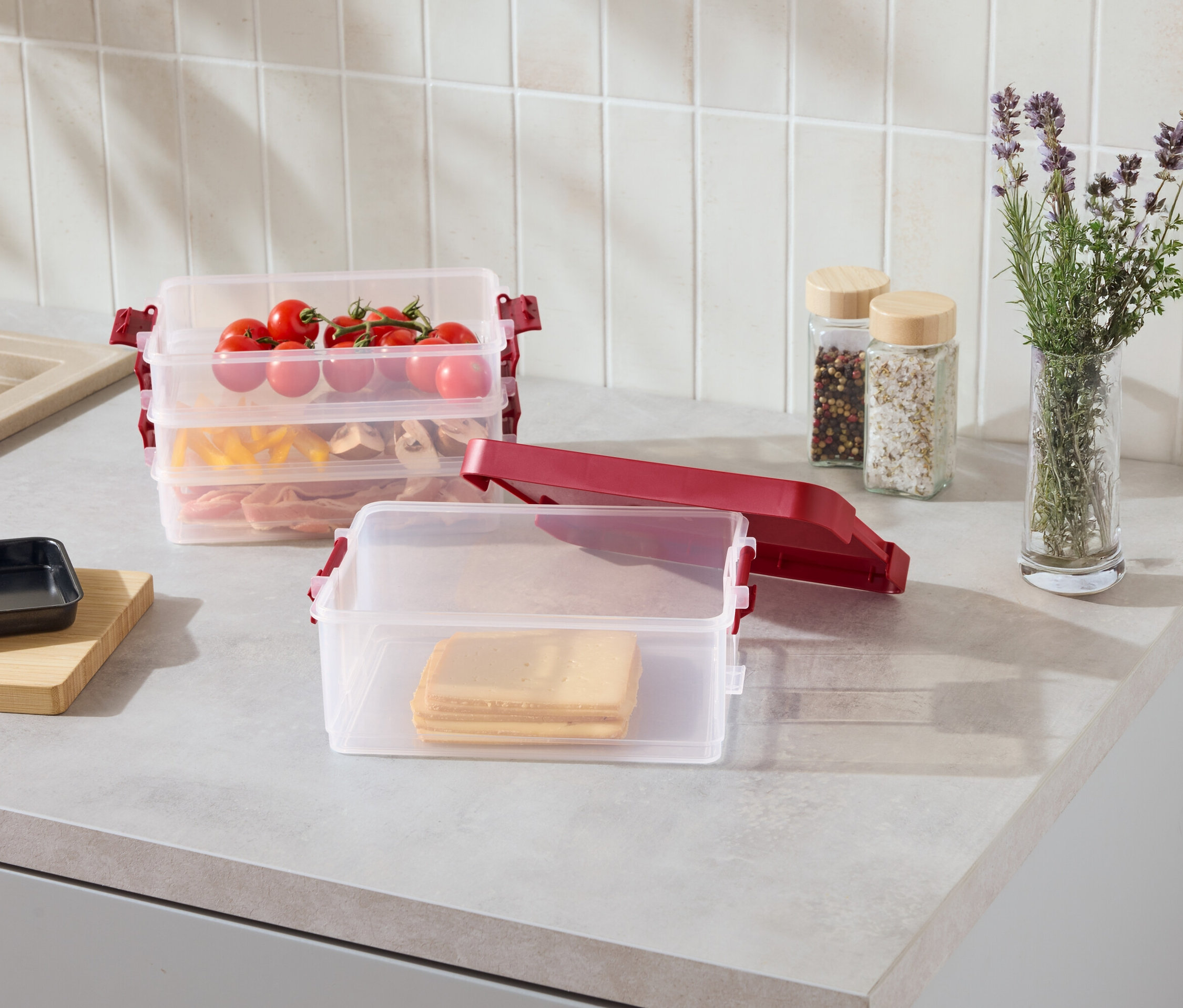 Na lince jsou průhledné stohovatelné nádoby s červenými sponami. Jedna obsahuje plátky sýra, další těstoviny a maso, a horní rajčata.