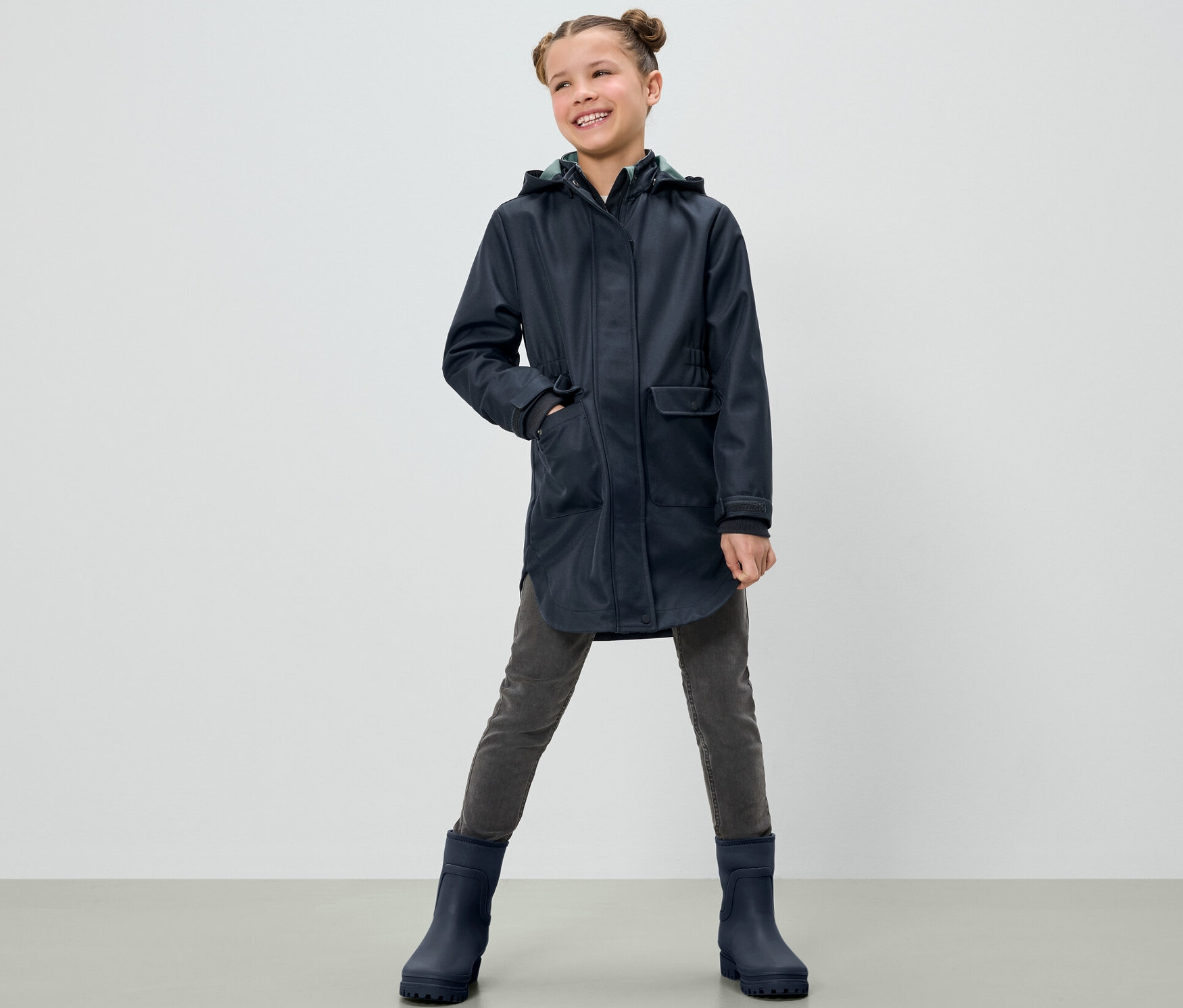 Usmívající se dívka pózuje v dětském softshellovém kabátu, šedých kalhotách a modrých botách.