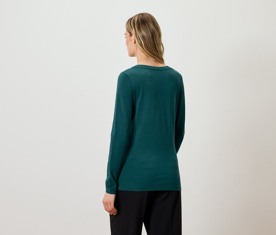 Zezadu je vidět žena s blond vlasy, která má na sobě zelené tričko s dlouhým rukávem a černé kalhoty.