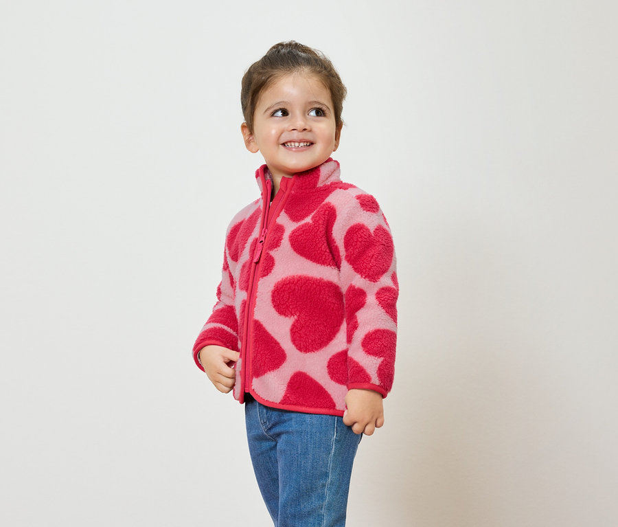 Dívka v růžové fleecové bundě se vzorem srdcí a modrých džínách se usmívá a dívá se stranou.