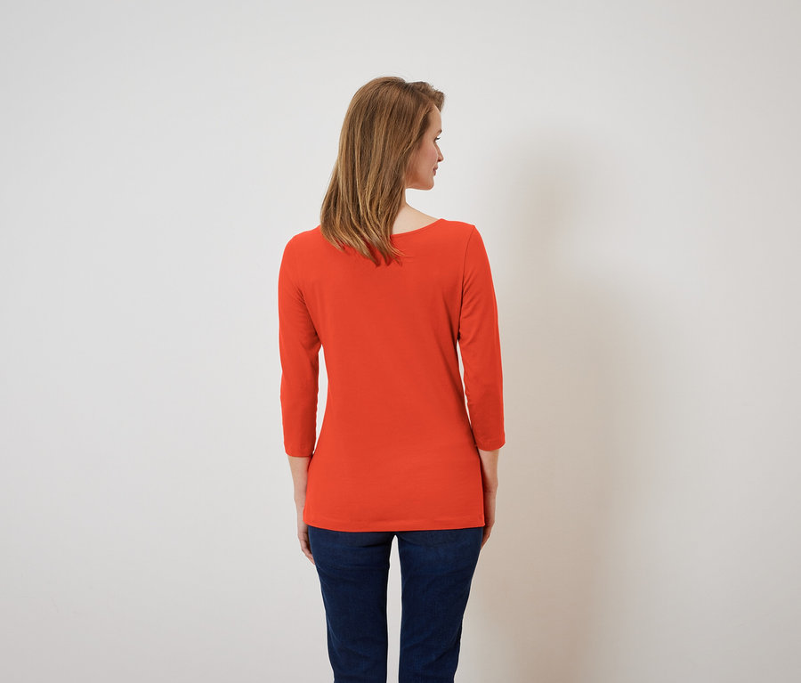 Žena zády k fotoaparátu má na sobě oranžové tričko s tříčtvrtečními rukávy a tmavě modré džíny.